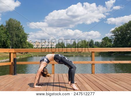Young Slim Girl In Grey Sport Wear Doing Yoga On The Bridge. Flexible Young Girl With Red Hair.