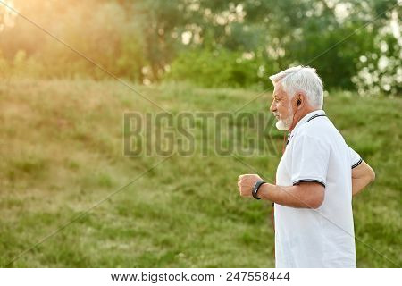 Old Man Running In Green City Park With Different Plants And Grass On Sunset. Wearing Classic White 