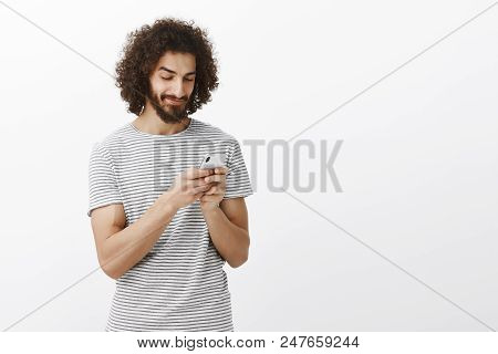 Studio Shot Of Intrigued Joyful Hot Hispanic Guy With Beard And Afro Haircut, Smiling At Screen Of S