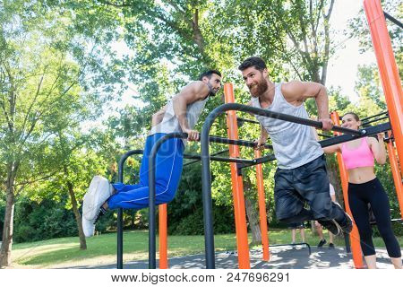 Two strong and motivated young men doing dips exercise for the upper body, during challenging workout with their friend in an outdoor fitness park with modern equipment