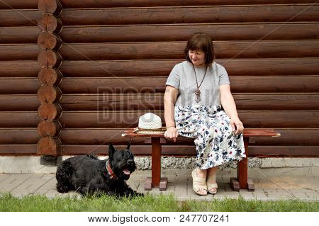 Middle-aged Woman In A Countryside Sitting On A Wooden Bench And Looking At A Black Schnauzer Near A