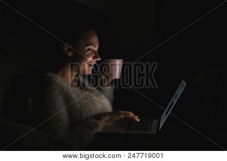 Cute Smiling Girl Sitting In Bed Late At Night, Looking Funny Ilm On Her Laptop And Drinking Tea
