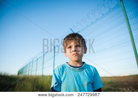 Cute Pensive Dirty Face Child Refugee Portrait On Fence With Razor Wire Bakdrop In Sunset