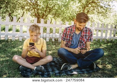 Concentrated Men Are Sitting On Blanket With Legs Crossed And Looking At Their Phones. They Are Play