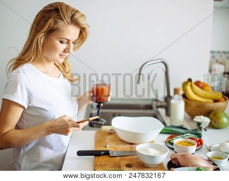 Smiling Caucasian Woman Holding Smoothie And Looking At Phone In The Domestic Kitchen Interior.