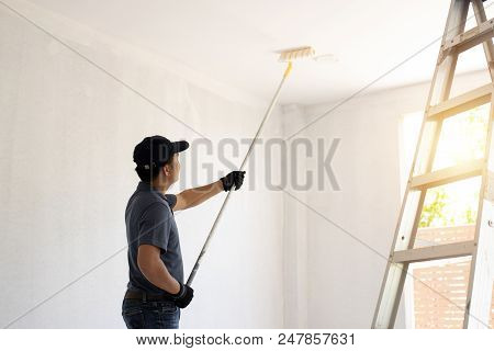 Asian Rear View Of A Male Painter Drawing A Wall With Paint Roller And A Separate Tank From A Large 