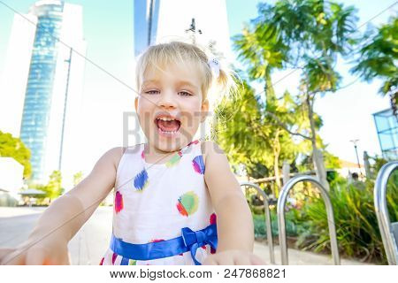 Close Up Portrait Of Emotional Cute Little Blondy Toddler Girl In Dress In The City Park With Modern