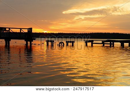 Wooden Pier Or Jetty Remains On Warm Lake Sunset And Sky Reflection Water.
