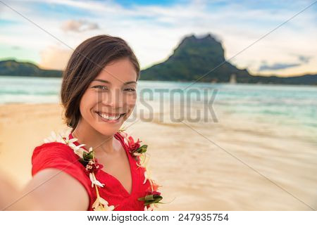 Luxury vacation selfie happy woman smiling taking photo with mobile phone on Bora bora island cruise travel destination at sunset. Smiling gorgeous Asian girl wearing Tahiti flower lei for luau.