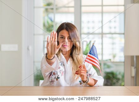 Young woman at home holding flag of united states of america with open hand doing stop sign with serious and confident expression, defense gesture