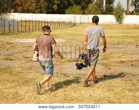 Bloggers Videographer And Photographer Go With The Camera On The Stabilizer On A Fenced Field With G