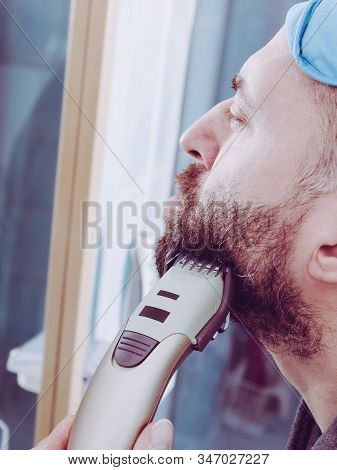 Focused Bearded Man Shaving His Beard Using Electric Razor. Taking Care Of Facial Hair, Wearing Slee