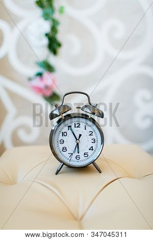 A Classic Metal Alarm Clock With A White Dial Stands On A Light Pouf On A Blurred Background.