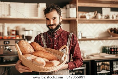 Bearded Hipster In Apron Carrying Wicker Basket With Loaves Of Fresh Bread During Work In Cozy Baker