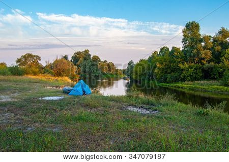 The Blue Tent Stands On The Bank Of The River Against The Background Of The Forest And The Blue Sky 