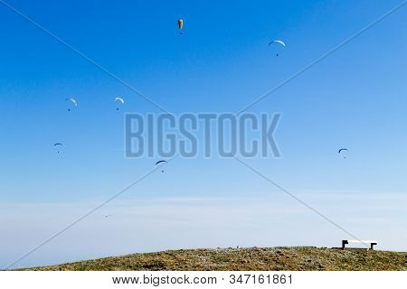 Paraglider On Blue Sky With Empty Bench On Foreground. Borso Del Grappa, Famous Paragliding Site, It