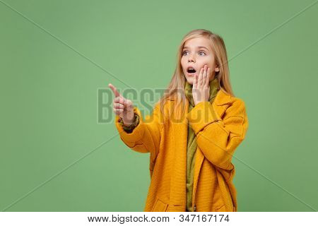 Shocked Little Blonde Kid Girl 12-13 Years Old In Yellow Coat Posing Isolated On Pastel Green Backgr