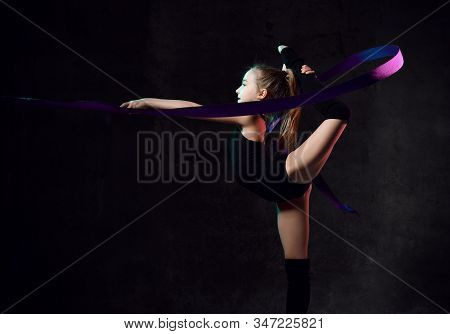 Young Girl Gymnast In Black Sport Body And Uppers Standing Sideways And Making Exercise With Blue Gy