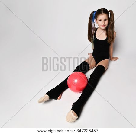 Cute Girl Gymnast With Long Hair In Black Sport Body And Uppers Sitting On Floor Holding Pink Gymnas