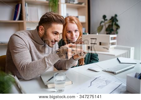 Architects With Model Of House Sitting At The Desk Indoors In Office, Working.