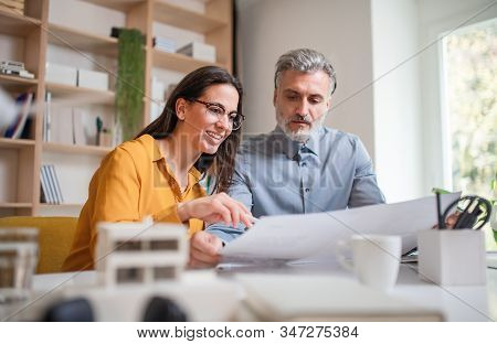 Architects With Model Of House Sitting At The Desk Indoors In Office, Working.