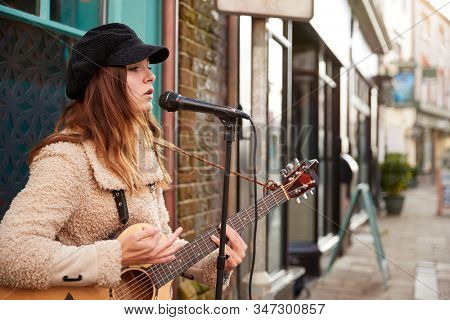 Female Musician Busking Playing Acoustic Guitar And Singing Outdoors In Street