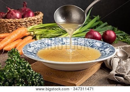 Pouring Chicken Bone Broth From A Ladle Into A Vintage Plate, With Fresh Vegetables In The Backgroun