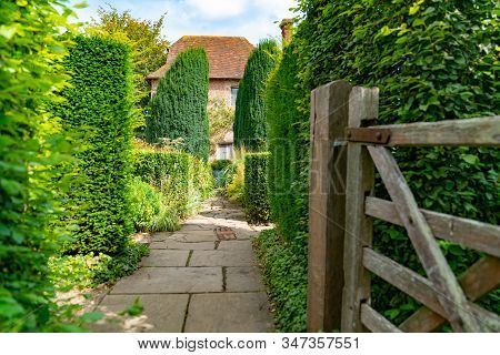 Garden Path Past Open Gate Between Trees And Plants With Building At End.
