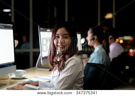 Asian young adult confidence operator woman agent with headsets working in a call center at night Environment with his colleague team as customer service. Using as Late night Hard working 24hr. Day. 