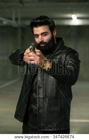 Merciless bearded assassin or terrorist in black leather jacket directing handgun on victim or rival before shooting