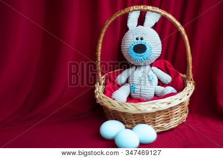Knitted Gray And Blue Toy Cute Easter Bunny In Willow Basket And Blue Dyed Eggs On Red Textile Backg