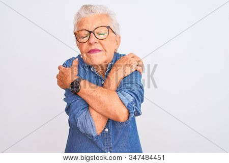 Senior grey-haired woman wearing denim shirt and glasses over isolated white background Hugging oneself happy and positive, smiling confident. Self love and self care