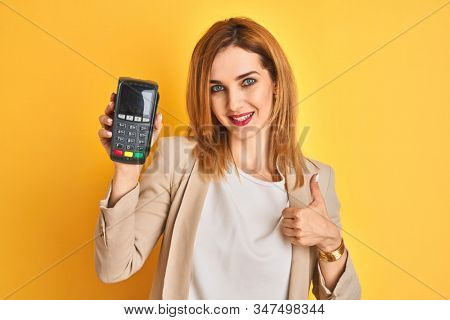 Redhead caucasian business woman holding point of sale terminal over isolated background happy with big smile doing ok sign, thumb up with fingers, excellent sign