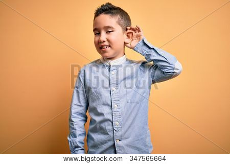 Young little boy kid wearing elegant shirt standing over yellow isolated background smiling with hand over ear listening an hearing to rumor or gossip. Deafness concept.