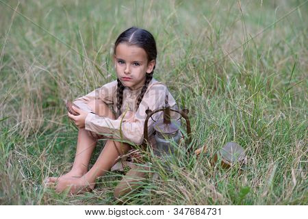 Pretty little girl with long pigtails and beautiful dirty face in summer forest and meadow holding vintage flask. Orphan, child of war, poverty, hunger, famine, poor destitute kid. 