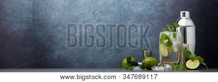 Set of bar tools: cocktail shaker, muddler, jigger, cocktail with ice in highball glass,  lime and mint on dark gray background with copy space. Bartender's tool. Mojito cocktail making.