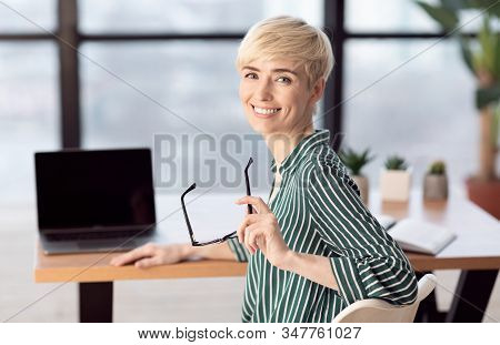 Happy Middle-aged Businesswoman Sitting At Laptop At Workdesk Smiling To Camera In Modern Office. Se