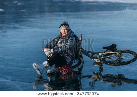 Woman Sitting Near Her Bike On Ice. The Girl Cyclist Stopped To Rest. She Sits On The Wheel And Enjo