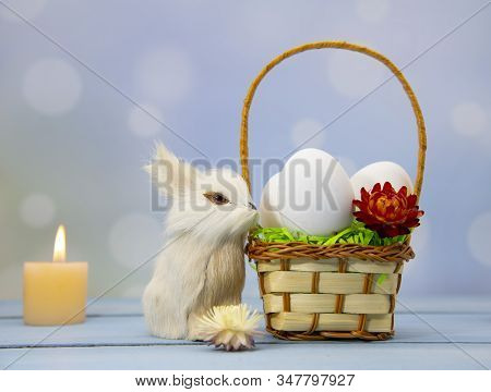 The Easter Bunny Sits Next To A Basket Full Of Eggs, Decorated With Dried Flowers. Burning Candles I