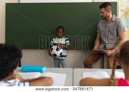 Front view of black schoolboy holding football and standing at green board in classroom of elementary school