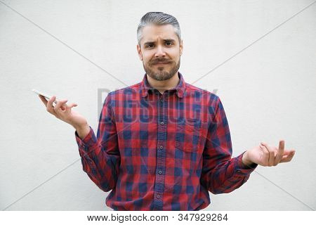 Bearded Young Man Holding Smartphone And Shrugging Shoulders. Stylish Guy Having Confused Look Facin
