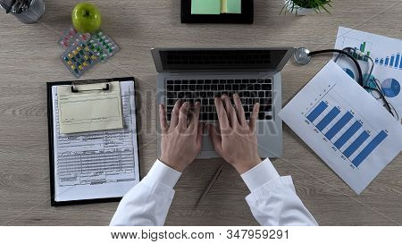 Physician Looking At Statistics Chart, Typing On Laptop, Preparing Presentation