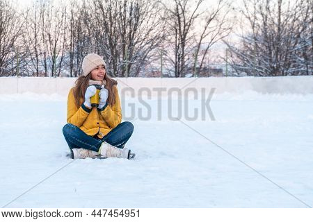 Portrait Of Young Woman In Ice Skates Sitting On The Snow, Drinking Coffee And Looking Away