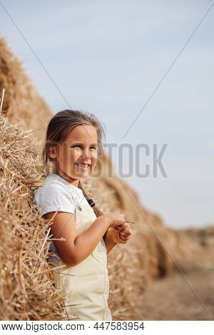 Blissful Young Female Child Standing Near High Haymow In Field With Braid On Shoulder, Hands Holding