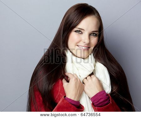 Portrait of a young smiling casual woman with long brunette hair on gray studio background, wearing red suede leather jacket and white scarf 