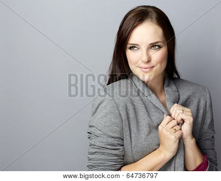 Portrait of a young smiling business woman with long brunette hair on gray studio background, wearing grey corporate blazer jacket 