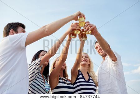 summer, holidays, tourism, drinks and people concept - group of smiling friends clinking bottles of beer or cider on beach