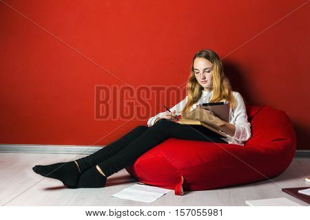 Young student girl studying sitting on the bag-chair near books and tablet pc