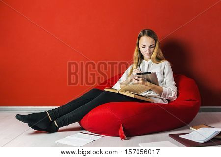 Young student girl studying sitting on the bag-chair near books and tablet pc
