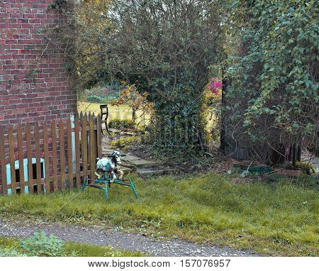 an old rustic metal rocking horse is in a green garden. Brick wall and timber fence behind